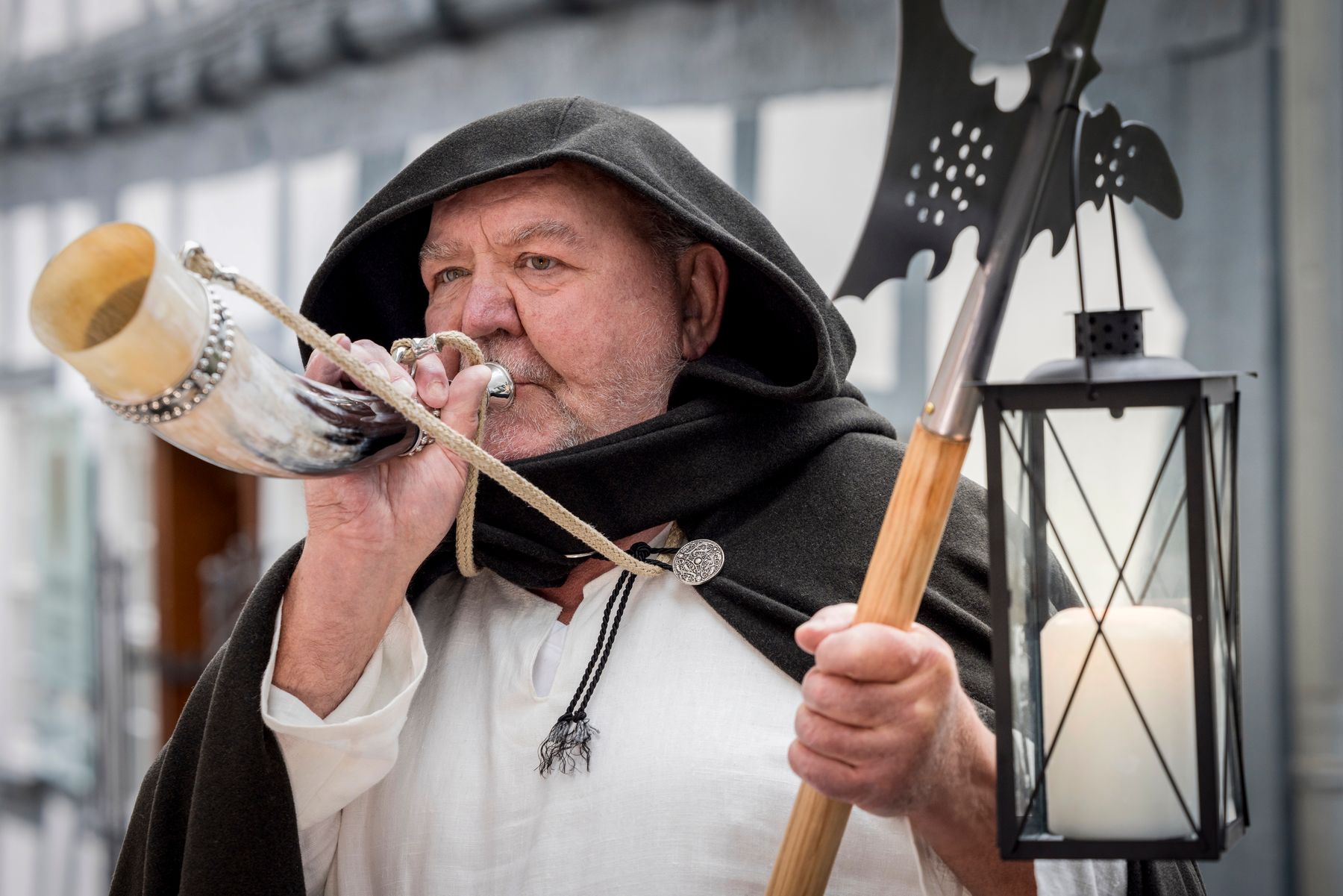 An older man in medieval costume blows a horn. He wears a black cloak with a hood, and holds a lantern and a poleaxe. A candle burns in the lantern, and traditional timbered buildings are visible in the background.